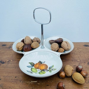 May include: A white ceramic three-section serving dish with a chrome stand. The dish has a citrus fruit design and is filled with nuts.