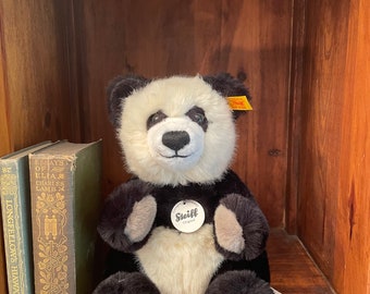 Könnte beinhalten: Ein schwarz-weißes Panda-Plüschtier, das auf einem Holzregal sitzt. Der Panda trägt ein Steiff-Etikett und hat eine schwarze Nase und Augen. Der Panda sitzt neben zwei Büchern.