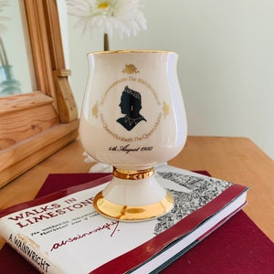 Puede incluir: Un gobelet conmemorativo de cerámica blanca con ribetes dorados, que presenta una silueta negra de la reina Isabel, la reina madre. El gobelet conmemora su 80 cumpleaños el 4 de agosto de 1980.