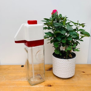 Puede incluir: Un dispensador de vidrio blanco y rojo con una tapa roja y un tubo de vidrio transparente. El dispensador está sentado sobre una superficie de madera junto a una planta en maceta con flores rosas.