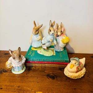 Puede incluir: Una colección de figuras de cerámica con personajes de los cuentos de Beatrix Potter. Las figuras incluyen conejos, un ratón y un pato, todos con trajes coloridos y detallados. Las figuras están dispuestas con títulos de libros antiguos visibles.