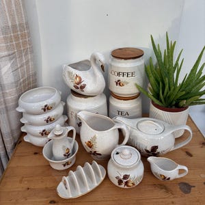 May include: A collection of vintage ceramic kitchenware with a floral design. Includes a teapot, creamer, sugar bowl, canisters labeled "Coffee" and "Tea", a swan-shaped planter, and a toast rack. The pieces are cream-colored with brown and yellow floral accents.