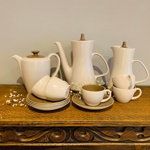 Puede incluir: Un juego de tazas y platillos de té de cerámica blanca con detalles marrones, junto con dos teteras a juego. El juego está dispuesto sobre una superficie de madera.