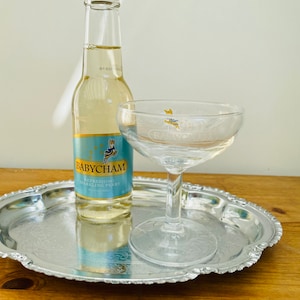 Puede incluir: Una botella de vidrio transparente de Babycham sidra espumosa en una bandeja plateada con una copa de champán de vidrio transparente.