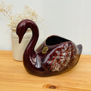 Puede incluir: Un macetero de cerámica en forma de cisne, de color marrón glaseado con un acabado texturizado. El macetero tiene una etiqueta que dice "Forever Yours, Made in England, Handpainted, Ceramic".