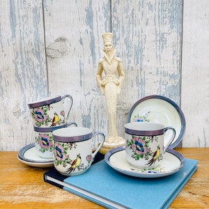 May include: Set of three vintage teacups and saucers featuring a floral and bird design. The cups and saucers are white with a blue and purple rim. The floral design includes pink, yellow, and blue flowers with yellow birds.