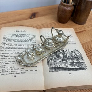 Puede incluir: Un juego de té de plata con una tetera, una jarra de crema y un azucarero en una bandeja de plata. El juego de té es del libro Alicia en el País de las Maravillas y presenta una escena de Alicia en una fiesta del té con el Sombrerero Loco, la Liebre de Marzo y el Lirón.