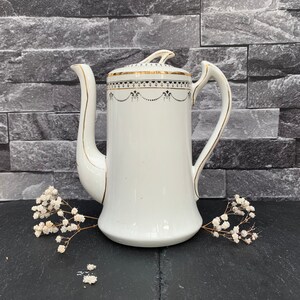 May include: White porcelain coffee pot with a gold trim and black floral design. The pot has a spout and a handle.