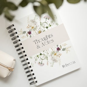 Könnte beinhalten: Ein weißes Notizbuch mit Spiralbindung und einem floralen Design. Auf dem Cover steht "Thoughts & Notes". In der unteren rechten Ecke des Covers steht der Name "Rebecca".