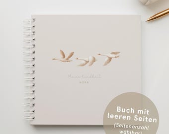 May include: A cream-coloured spiral-bound notebook with an illustration of three flying swans. The text "Meine kindheit" and the name "NORA" are printed below the swans. A cream pencil case and a gold pen are also visible.
