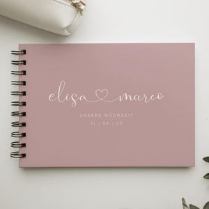 Puede incluir: Un cuaderno en espiral rosa empolvado con los nombres "Elisa & Marco" en escritura elegante, un símbolo de corazón y la fecha "01 | 08 | 25". El cuaderno está sobre una superficie blanca, con una bolsa blanca y vegetación en el fondo.
