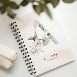 Könnte beinhalten: Ein weißes Notizbuch mit Spiralbindung und einem rosafarbenen Aquarelldesign sowie dem Text "Annikas Notizbuch" auf dem Einband.