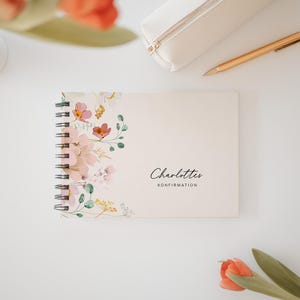 Könnte beinhalten: Ein cremefarbenes Spiralnotizbuch mit floralem Design und dem Text "Charlottes Konfirmation" in schwarzer Schrift. Ein goldener Stift und ein weißes Federmäppchen liegen daneben. Das Notizbuch hat ein florales Muster.