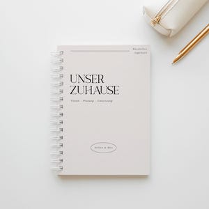 Könnte beinhalten: Ein cremefarbenes Spiralnotizbuch mit dem Text "UNSER ZUHAUSE" in Schwarz und "Vision - Planung - Umsetzung" darunter. Ein weißes Federmäppchen und ein goldener Stift liegen auf der weißen Oberfläche. Das Notizbuch trägt auch den Text "Selina & Max".
