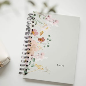 Könnte beinhalten: Ein weißes Spiralnotizbuch mit einem Blumendesign mit rosa, grünen und gelben Aquarellblumen. Das Notizbuch trägt den Namen "Laura" auf dem Einband.