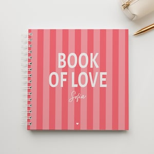 Puede incluir: Un cuaderno cuadrado de espiral con una cubierta de rayas verticales en rosa y rosa claro. El texto BOOK OF LOVE está impreso en letras mayúsculas blancas, seguido del nombre Sofia en letra cursiva blanca y un pequeño corazón blanco.
