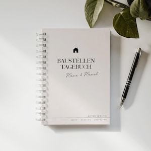 Könnte beinhalten: Ein weißes Spiralnotizbuch mit dem Text "BAUSTELLEN TAGEBUCH Marie & Marcel" und einem kleinen Haussymbol. Ein schwarzer und silberner Stift liegt neben dem Notizbuch, mit grünen Blättern im Hintergrund. Das Notizbuch ist für die Renovierungsplanung.