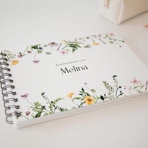 Könnte beinhalten: Ein weißes Spiralnotizbuch mit floralen Aquarellillustrationen in Gelb, Rosa und Lila. Der Text "Konfirmation von Melina" ist in Schwarz gedruckt. Ein Bleistift und ein weißes Federmäppchen befinden sich im Hintergrund.