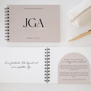 Könnte beinhalten: Ein spiralgebundenes Notizbuch mit hellbraunem Einband, mit dem Text "ERINNERUNGSBUCH" und "JGA Selina BRIDE TO BE." Ein offenes Notizbuch zeigt handschriftlichen Text. Ein weißes Federmäppchen und ein goldener Stift sind ebenfalls sichtbar.