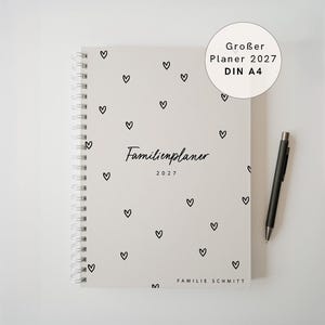 Könnte beinhalten: Ein hellgrauer Spiralplaner mit einem Muster aus kleinen schwarzen Herzen. Auf dem Einband steht "Familienplaner 2027" und unten "FAMILIE SCHMITT". Ein runder Aufkleber zeigt "Großer Planer 2027 DIN A4". Ein schwarzer Stift liegt daneben.