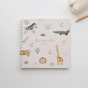 Könnte beinhalten: Ein spiralgebundenes Notizbuch mit hellrosa Einband mit Aquarellillustrationen eines Zebras, einer Maus, eines Krokodils, eines Tigers, einer Giraffe und eines Igels. Das Notizbuch trägt den Text "Mein erstes Jahr JONTE" und kleine Herz- und Pflanzenmotive.