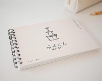 May include: White spiral bound journal with the text "Bride to be MARIE" and "MEIN JGA" printed on the cover. A black and white illustration of a champagne glass tower is above the text.