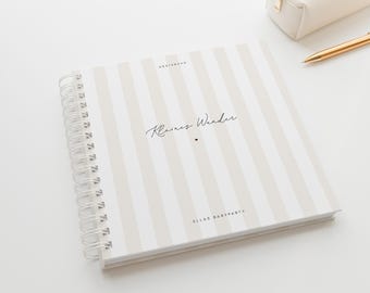 May include: A beige and white striped guest book with spiral binding. The cover features the words "Kleines Wunder" and "ELLAS BABYPARTY". A gold pen and a cream pencil case are visible in the background, on a white surface.