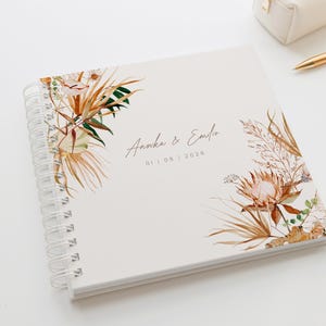 Puede incluir: Un cuaderno de espiral de color crema, adornado con diseños florales en acuarela en tonos marrón, verde y beige. Los nombres "Annika & Emilio" y la fecha "01 | 08 | 2026" están elegantemente escritos en la portada.