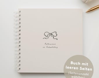 May include: A cream-coloured spiral-bound notebook with a black bow illustration and handwritten text. A small cream-coloured pouch and a gold pen are visible in the background.