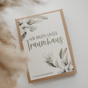 Könnte beinhalten: Eine rechteckige Box mit hellbraunem Karton außen. Die Box enthält eine weiße Karte mit dem Text "WIR BAUEN UNSER Traumhaus" und floralen Illustrationen. Der Text "40 MEILENSTEINKARTEN" ist ebenfalls sichtbar.