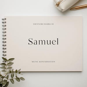 Könnte beinhalten: Ein cremefarbenes Spiralbuch mit dem Namen "Samuel" in Schwarz gedruckt. Der Text "ERINNERUNGSBUCH" und "MEINE KONFIRMATION" stehen über und unter dem Namen. Ein goldener Stift und ein cremefarbenes Federmäppchen sind im Hintergrund.
