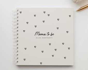 May include: A white spiral-bound notebook with a cover scattered with small black hearts. The words "Mama to be" and "ELLAS BABYPARTY" are written in black script. A gold pen and a small white pouch are in the background.