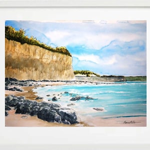 May include: Watercolour painting of a coastal scene, framed in white. The artwork depicts a light brown cliff with green foliage, a sandy beach, and turquoise water under a blue sky with clouds. Dark rocks line the shore.