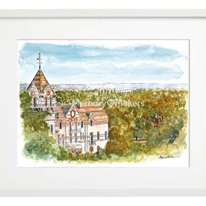 May include: Watercolour painting of two ornate buildings with red tile roofs and white walls, set amidst lush green trees. The artwork is framed in a white mount and frame, with a blue sky backdrop.