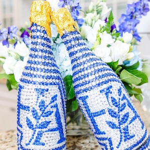 May include: Two champagne bottles decorated with blue and clear crystals. The bottles have a blue and white design with a floral motif. The bottle tops are covered in gold foil, and the background features a bouquet of white and blue flowers.