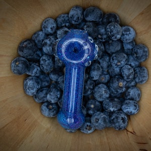 May include: A blue glass pipe with glitter embedded in the glass. The pipe is sitting on a wooden surface surrounded by blueberries.