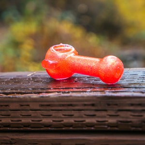 May include: A red glass pipe with a sparkly finish sits on a wooden surface.