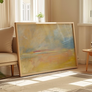 May include: A large, framed abstract painting with a light wood frame. The artwork features a blend of yellow, blue, and red hues, creating a soft, atmospheric landscape. The painting leans against a wall in a well-lit living room.