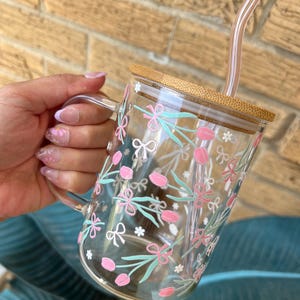 May include: A clear glass mug with a floral design featuring pink flowers, green leaves, and white bows. The mug has a bamboo lid and a clear glass straw.