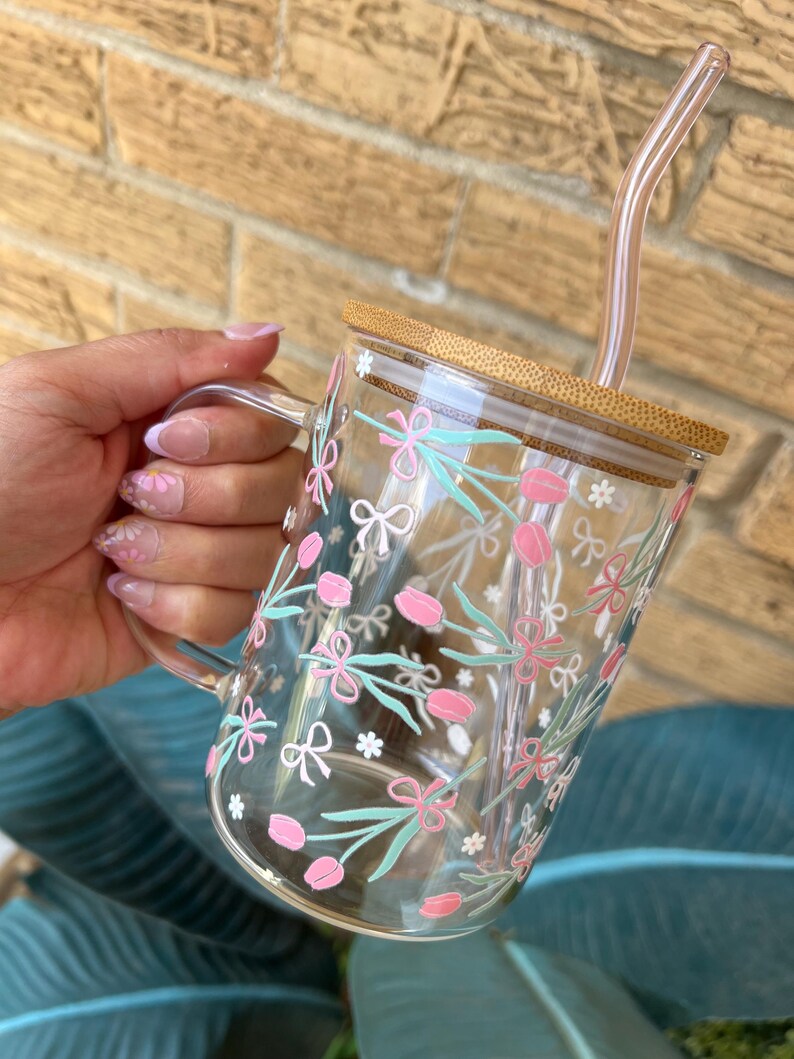 May include: A clear glass mug with a floral design featuring pink flowers, green leaves, and white bows. The mug has a bamboo lid and a clear glass straw.