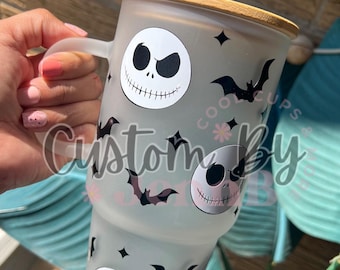 May include: A frosted glass tumbler with a black and white Jack Skellington design and black bats. The tumbler has a wooden lid and a straw.