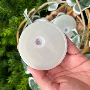 May include: A white plastic lid with a small hole in the center. The lid is smooth and has a slightly raised edge.