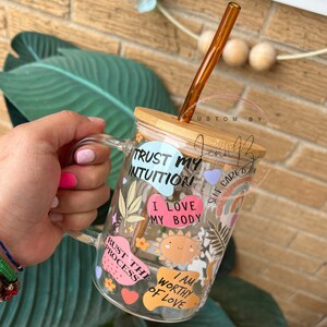 May include: A clear glass mug with a bamboo lid and a brown straw. The mug has various colorful stickers with positive affirmations, including "Trust My Intuition", "I Love My Body", "Trust The Process", and "I Am Worthy Of Love".