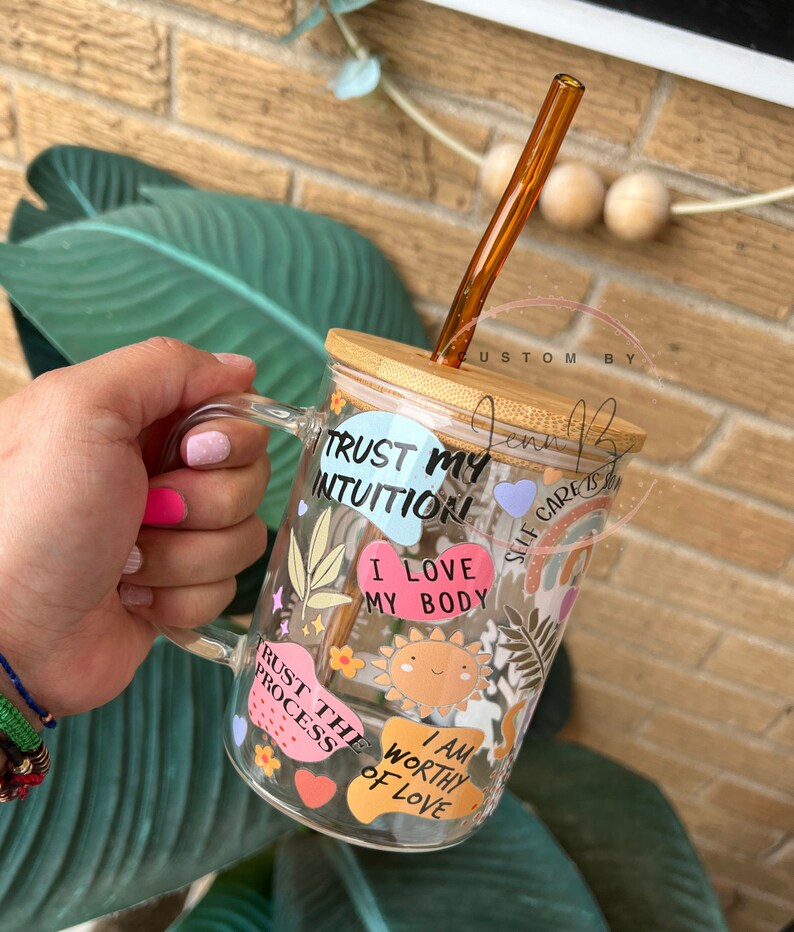May include: A clear glass mug with a bamboo lid and a brown straw. The mug has various colorful stickers with positive affirmations, including "Trust My Intuition", "I Love My Body", "Trust The Process", and "I Am Worthy Of Love".