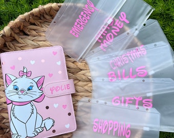 May include: A pink and white polka dot binder with a cartoon cat illustration and the word "Polie" on the front. The binder is surrounded by clear plastic zippered pouches with pink lettering. The pouches are labeled "Emergency", "This Money", "Christmas Bills", "Gifts", and "Shopping".