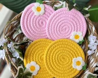 May include: Four round woven coasters with daisy embellishments. Two coasters are pink and two are yellow.
