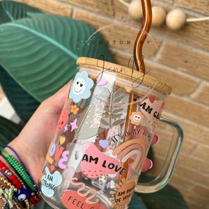 May include: A clear glass mug with a bamboo lid and a brown glass straw. The mug is decorated with colorful stickers that say "I am love", "I am strong", "I feel calm", "I am proud of myself", and "It's okay to not be okay".