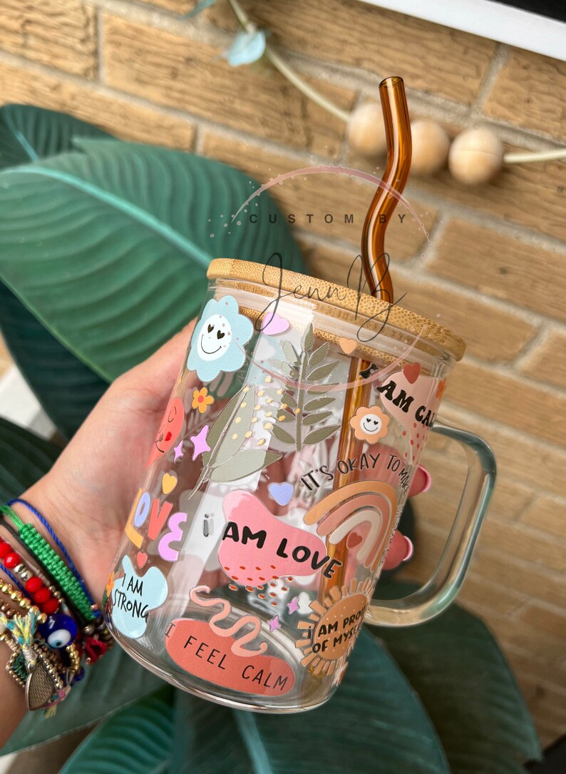 May include: A clear glass mug with a bamboo lid and a brown glass straw. The mug is decorated with colorful stickers that say "I am love", "I am strong", "I feel calm", "I am proud of myself", and "It's okay to not be okay".