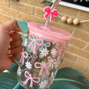 May include: A pink glass mug with a pink lid and a pink straw with a bow on top. The mug has a white and pink floral design with bows.