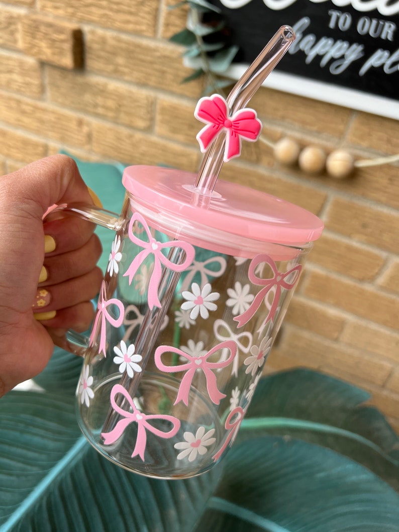 May include: A pink glass mug with a pink lid and a pink straw with a bow on top. The mug has a white and pink floral design with bows.
